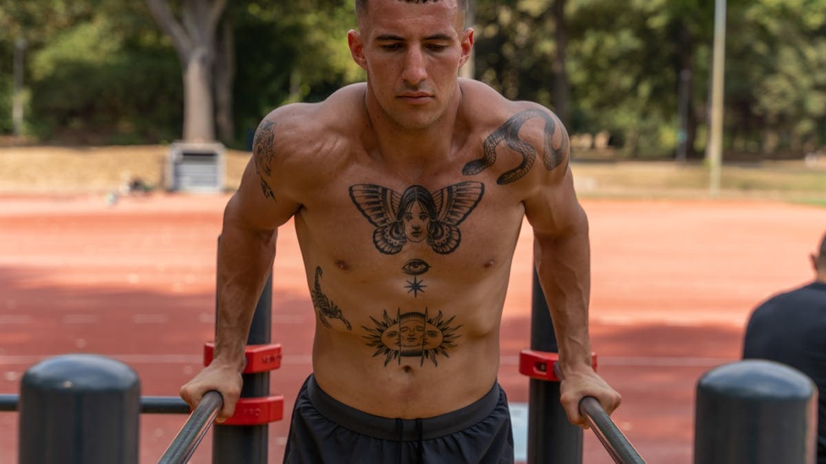 Strong shirtless man with tattoos performing outdoor workout on parallel bars in summer.